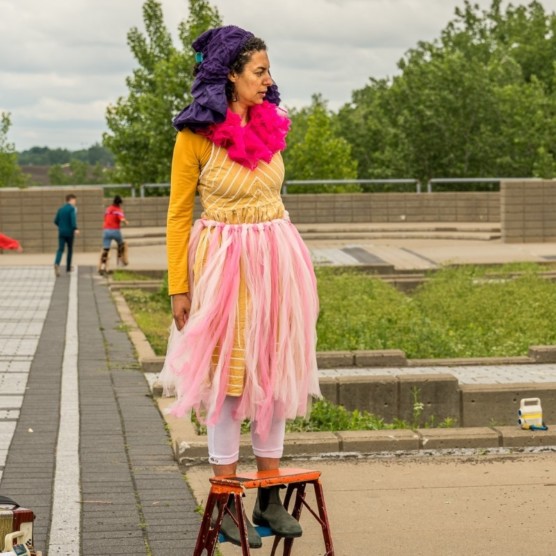 Photo en extérieur du personnage d'Amarelinha debout sur un escabeau. Elle porte un habit jaune, une jupe en tulle rose, un foulard fuchsia et un tissus mauve foncé sur la tête. Sa tête est tournée vers la droite et elle regarde devant elle.