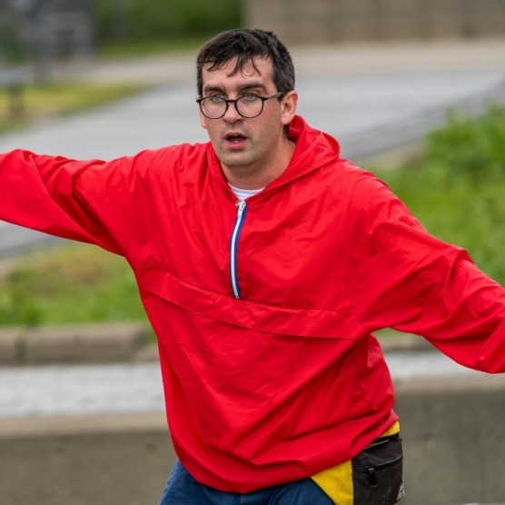Photo en extérieur du personnage de Sam, portant un coupe-vent rouge et des lunettes à monture noire. Il a les bras ouverts et regarde devant lui, la bouche ouverte.