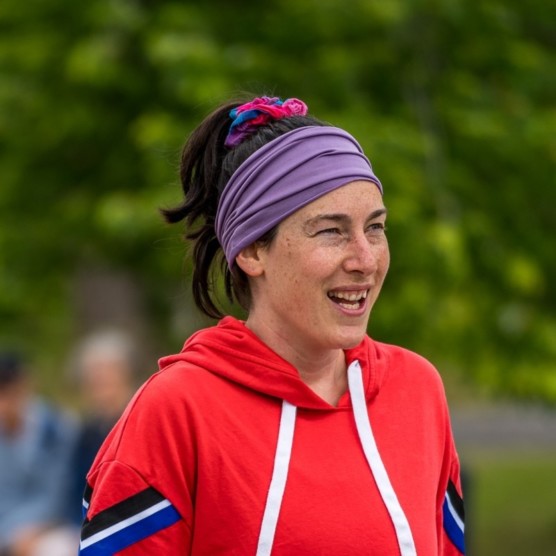 Photo en extérieur du personnage de Simone, qui porte un coton ouaté rouge et un foulard mauve dans ses cheveux attachés. Elle est légèrement tournée vers la droite et regarde devant elle les yeux plissés, en souriant.