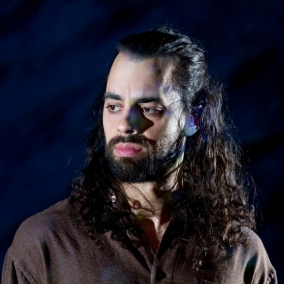 Portrait de Solo, un homme au teint moyen-clair, aux yeux bruns, aux longs cheveux bruns ondulés avec une barbe brune. Il porte une chemise brun pâle, son visage est tourné vers la lumière à gauche et il regarde devant lui. Le fond est bleu et noir.
