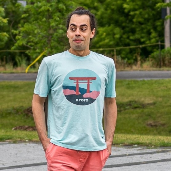 Photo du personnage d'Ulysse, les cheveux attachés, il porte un t-shirt aqua avec un dessin de Kyoto et des shorts couleur saumon. Les mains dans les poches, il regarde vers la gauche en souriant, les lèvres pincées.