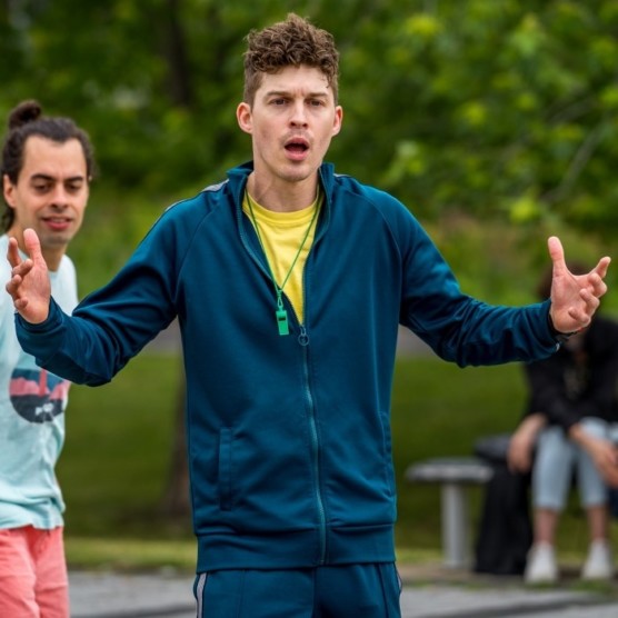 Photo en extérieur du personnage de Victor, qui porte un habit de sport bleu, un t-shirt jaune et un sifflet autour du cou. Ses bras sont ouverts et il a un visage expressif, la bouche ouverte.