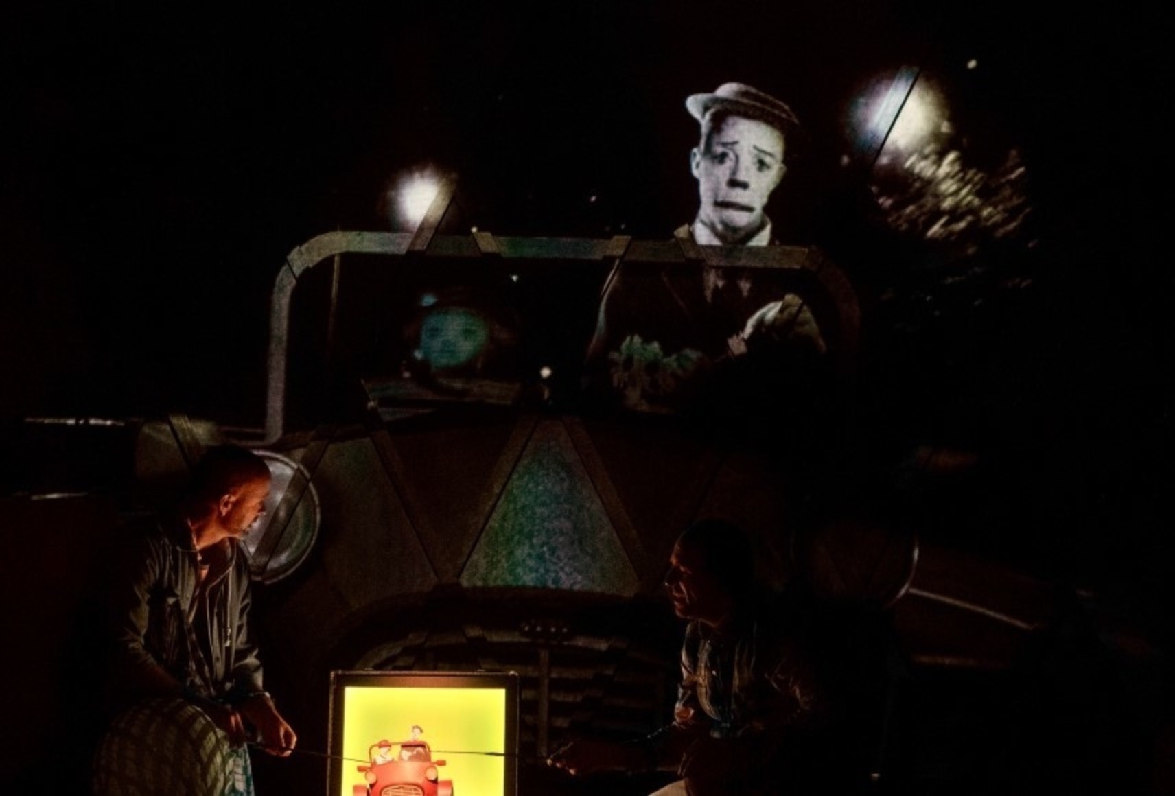 Photo des comédiens jouant avec une maquette de voiture devant des projections vidéo