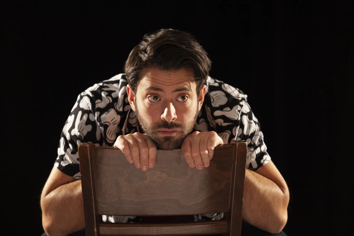 Photo du comédien sous un éclairage latéral, devant un fond noir. Il porte un t-shirt brun et une chemise à manches courtes aux motifs floraux. Il est appuyé contre un dossier de chaise en bois, qu'il tient des deux mains. Il regarde devant lui.