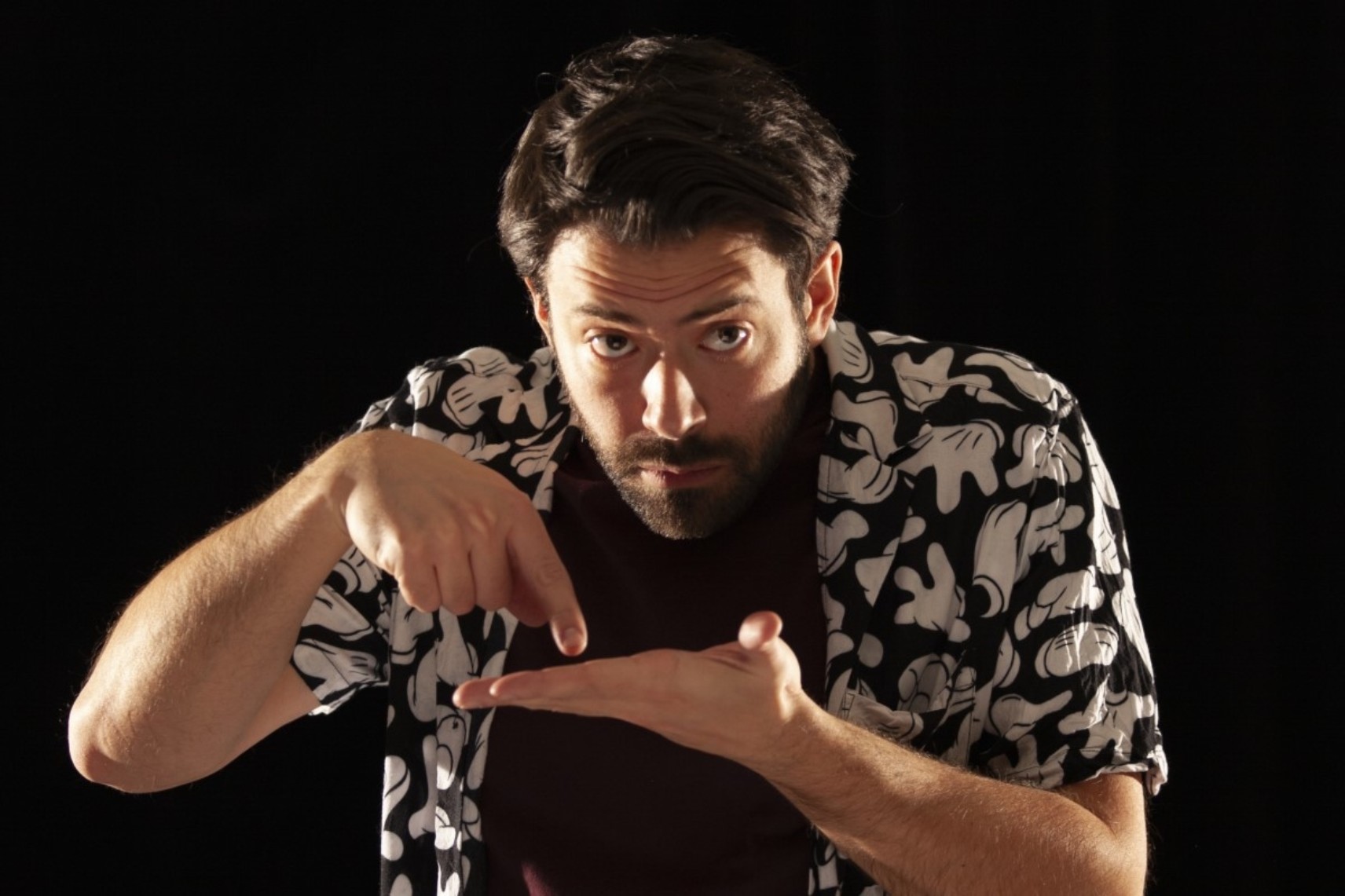 Photo du comédien sous un éclairage latéral, devant un fond noir. Il porte un t-shirt brun et une chemise à manches courtes aux motifs floraux. Sa main gauche est ouverte et l'index de sa main droite touche sa paume. Il regarde devant lui.