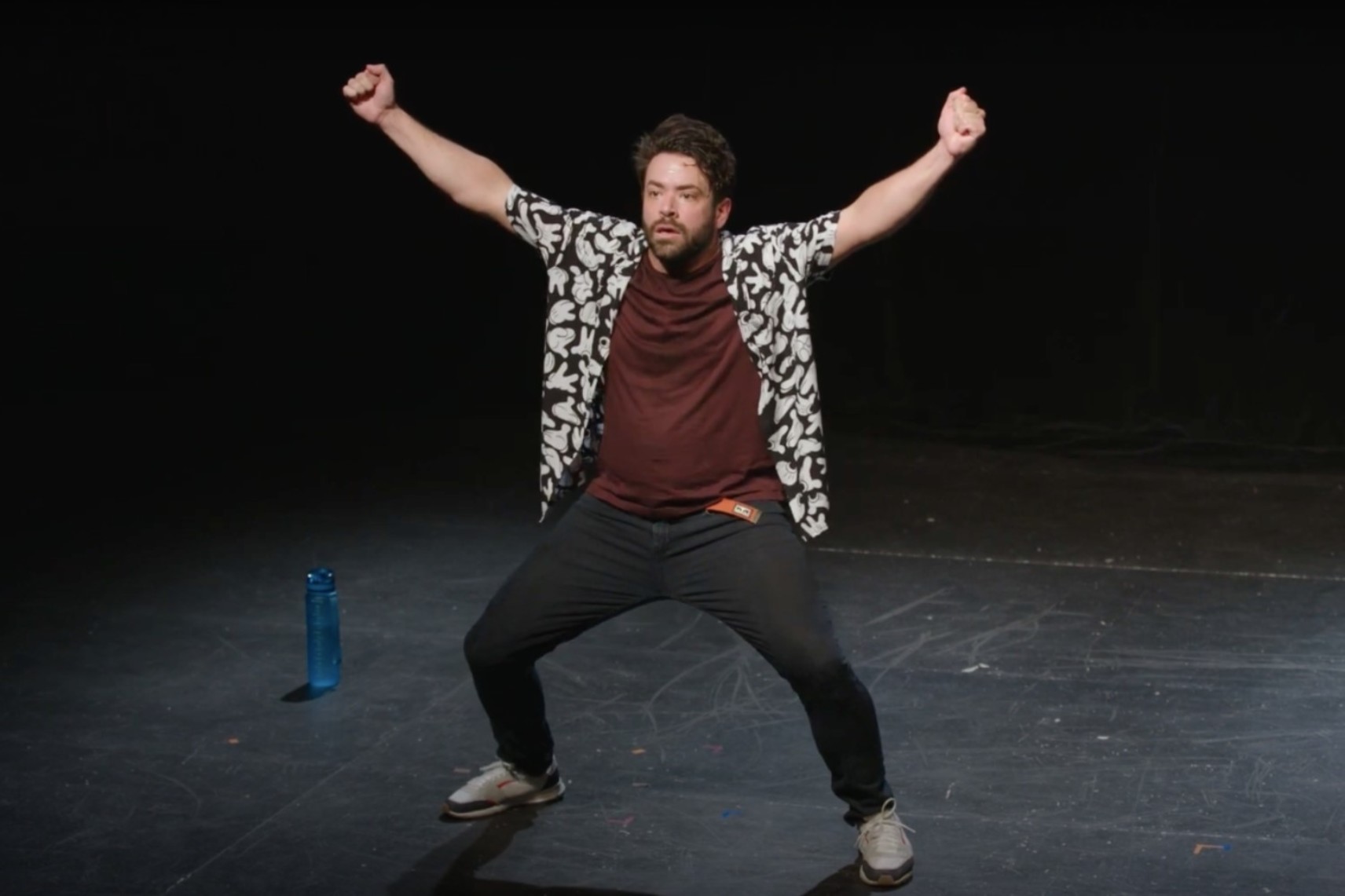 Un comédien est debout sur scène, les jambes écartées et les bras ouverts dans les airs, poings fermés. Il porte des pantalons noirs, un t-shirt brun et une chemise à manches courtes aux motifs floraux. L'arrière-plan est noir.