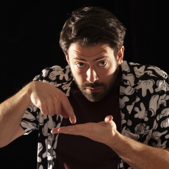 Photo du comédien sous un éclairage latéral, devant un fond noir. Il porte un t-shirt brun et une chemise à manches courtes aux motifs floraux. Sa main gauche est ouverte et l'index de sa main droite touche sa paume. Il regarde devant lui.