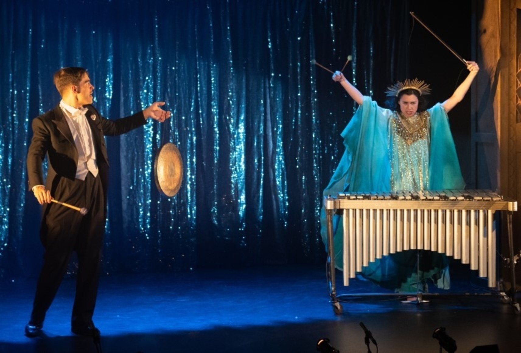 Dans un éclairage bleuté, un homme en tuxedo noir et blanc tient un petit gong de sa main gauche. En face de lui, une femme devant un xylophone, les bras dans les airs. Elle porte une large robe bleue et or avec bijou de tête ornemental dorée.