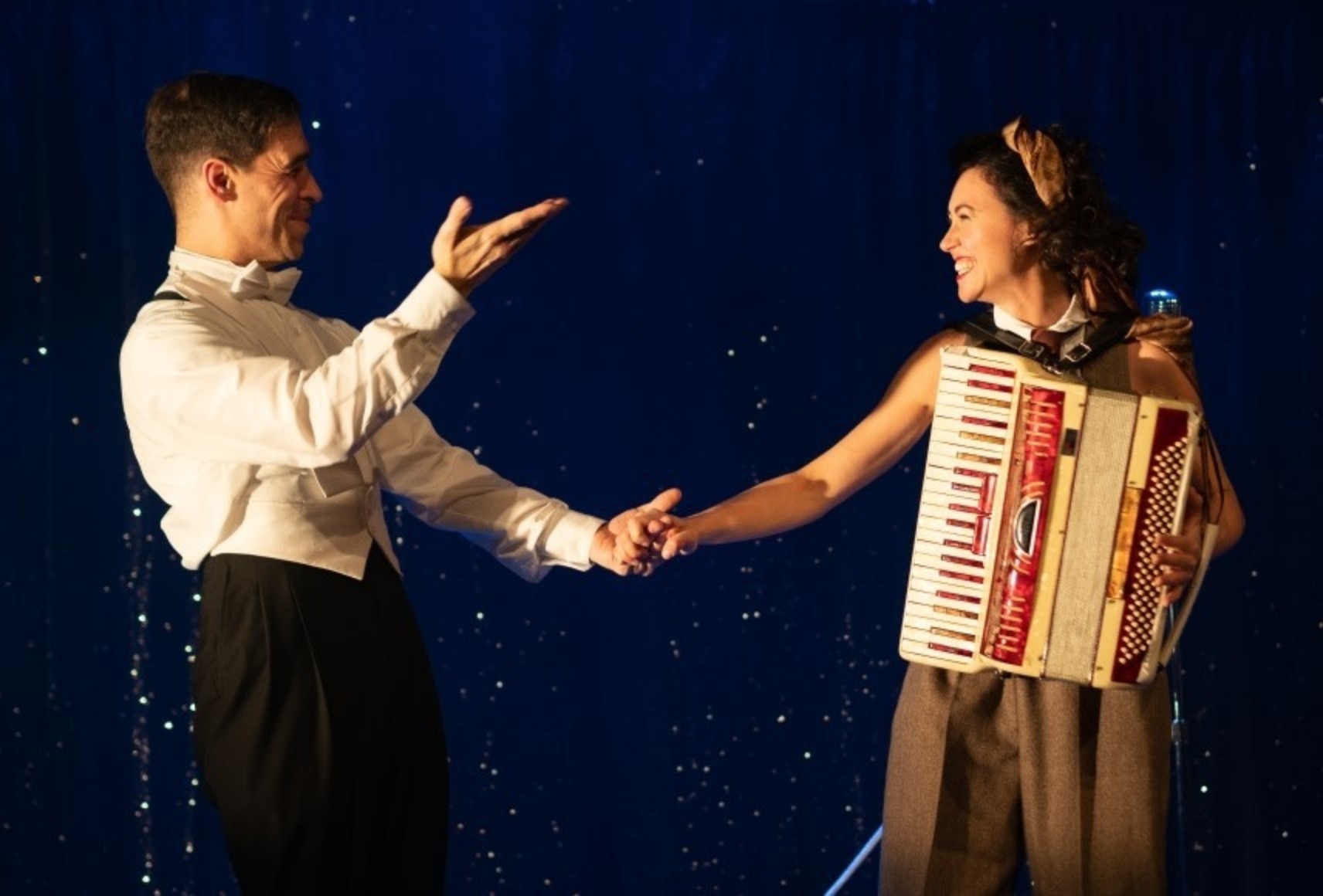 Un homme portant un pantalon noir et une chemise blanche tient la main d'une femme portant une pantalon brun et un accordéon sur son buste. Les deux se regardent et sourient.