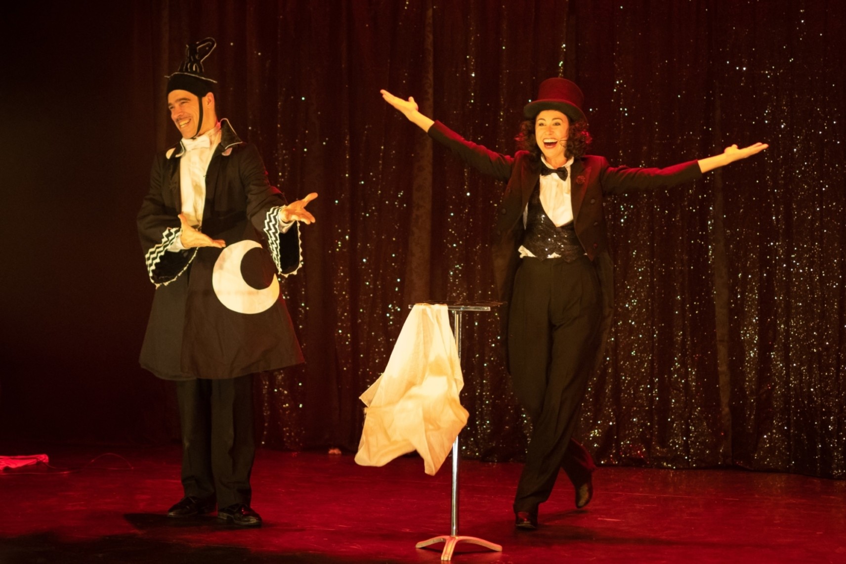 Deux personnages devant un rideau noir scintillant. À gauche, un homme avec un costume noir avec une lune blanche brodée. À gauche, une femme en tuxedo avec un haut de forme, les bras ouverts. Les deux sourient en regardant le public.