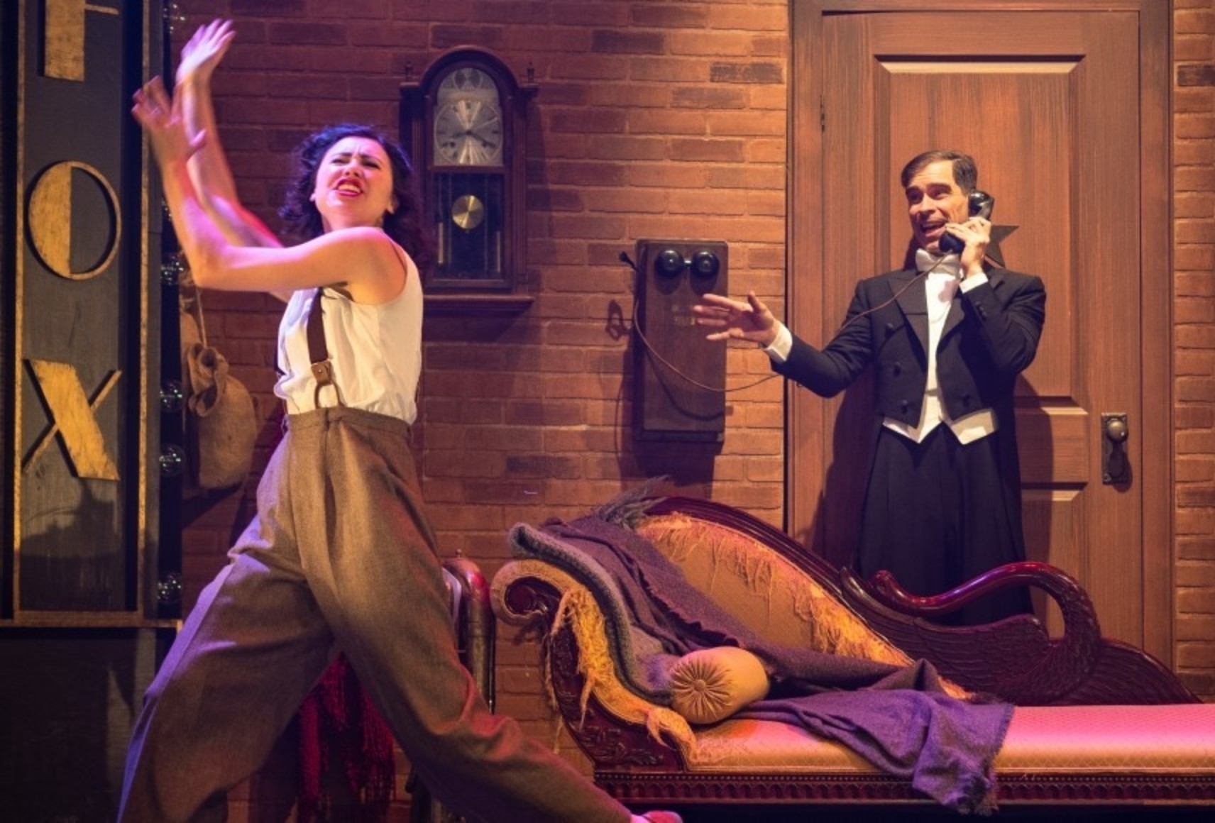Deux personnages dans un décor chargé. À gauche, une femme portant des pantalons bruns et un haut blanc dans une pose expressive. Derrière, à droite, un homme en tuxedo noir parlant au téléphone.