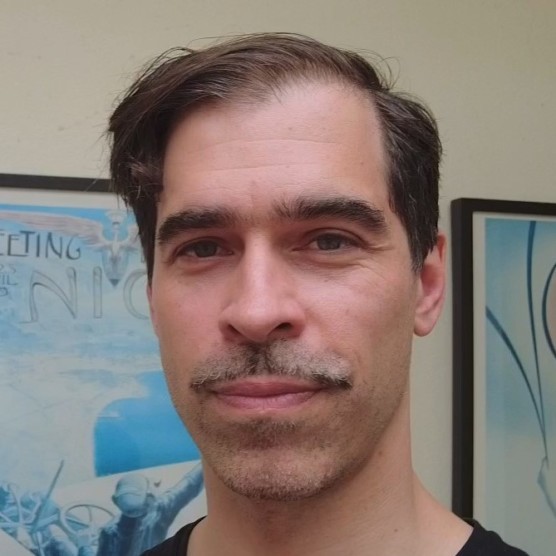 Photo de Marc Alexandre, un homme au teint clair, aux yeux bruns, aux cheveux bruns courts coiffés sur le côté gauche et à la moustache courte. Il regarde la caméra, le visage détendu. Derrière lui, des illustrations encadrées.