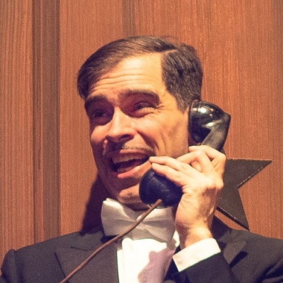 Photo de Marc Alexandre, portant un tuxedo noir et blanc, tenant l'acoustique d'un vieux téléphone noir à son oreille. Il a un visage expressif et regarde vers la gauche.