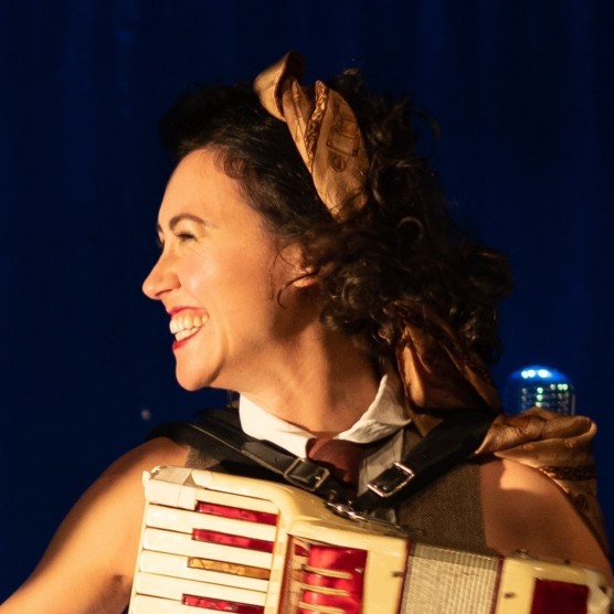 Photo du personnage de Marie de profil vers la gauche, les cheveux bruns et tenant un accordéon. Elle sourit en regardant devant elle.