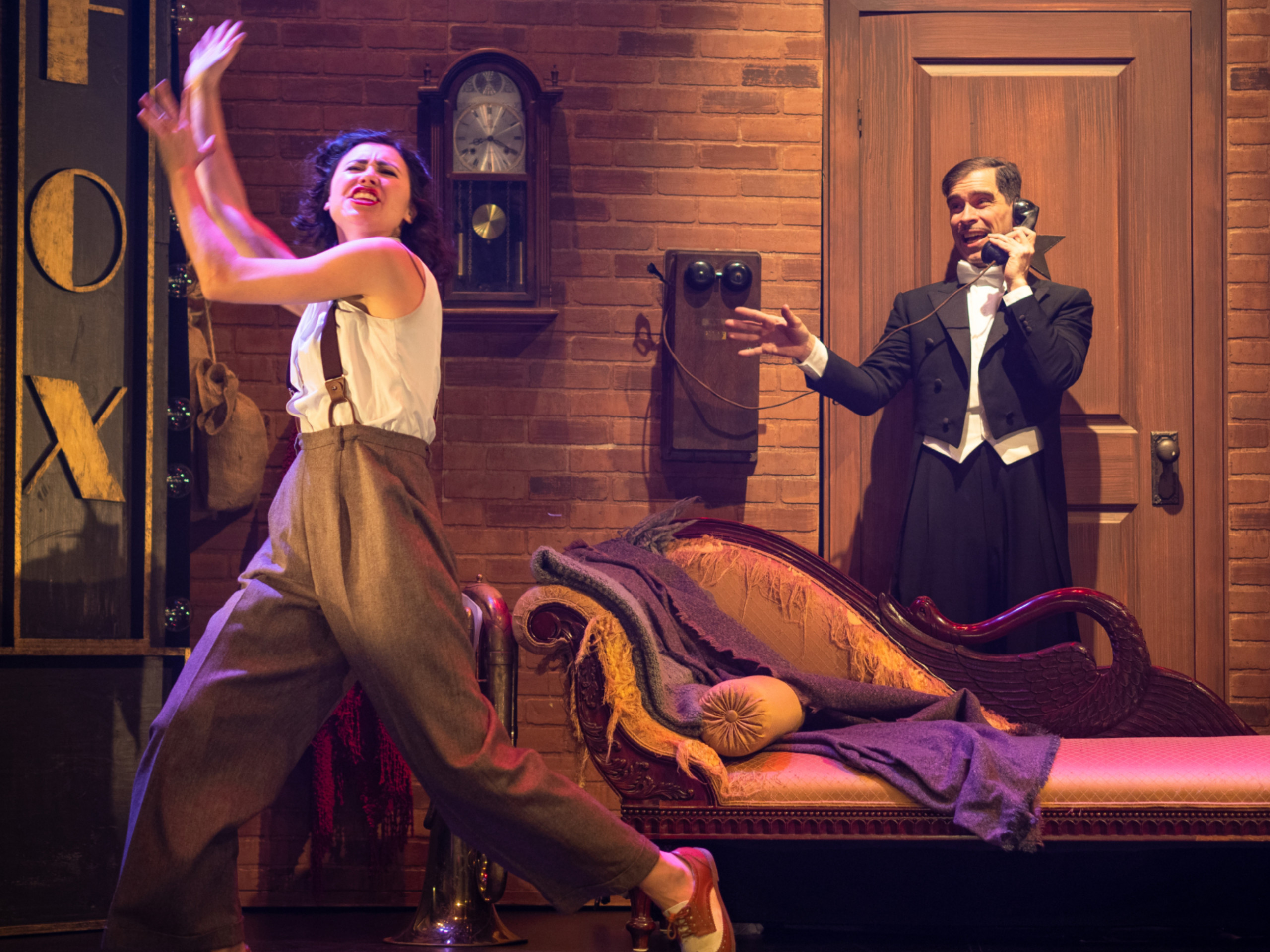 Deux personnages dans un décor chargé. À gauche, une femme portant des pantalons bruns et un haut blanc dans une pose expressive. Derrière, à droite, un homme en tuxedo noir parlant au téléphone.