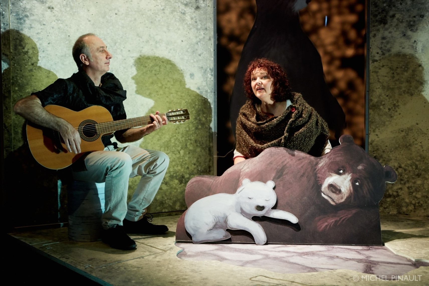À gauche, Christophe joue de la guitare tandis que Jasmine est derrière une grande figure d'ours brun avec un petit ourson blanc qui dort collé à son flanc. Leurs ombres sont projetées sur le décor.