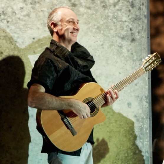 Photo de Christophe sur scène, jouant de la guitare. Il porte un haut noir au large col et des pantalons clairs. Son visage est tourné vers la droite et il sourit.