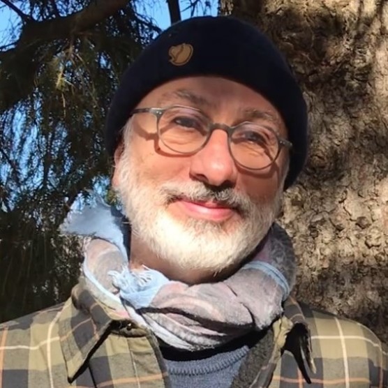 Photo de Christophe, un homme au teint clair, à la barbe blanche et portant des lunettes rondes. Il porte une tuque noire, un foulard bleuté, une chemise à carreaux vert kaki et il est photographié devant un arbre.