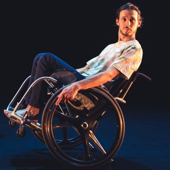 Portrait de l'artiste Jérémie Desbiens sur scène. On le voit de profil, assis dans un fauteuil roulant, incliné vers l'arrière. Il porte des pantalons bleu foncés et un haut pâle aux motifs tie dye. Son visage confiant est tourné vers le public.