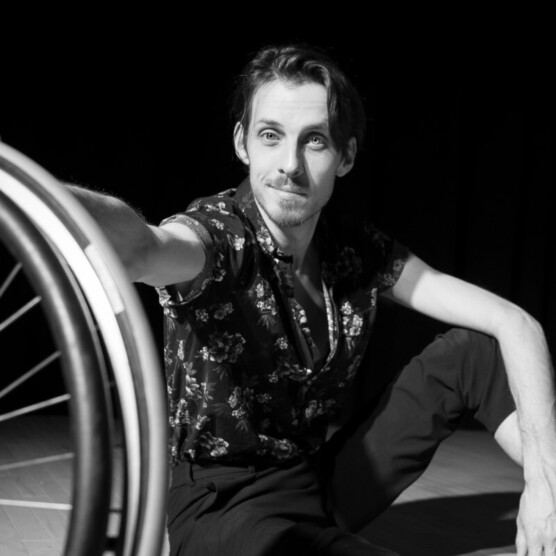 Portrait en noir et blanc de l'artiste Jérémie Desbiens. Il a les cheveux courts et est assis, le bras gauche appuyé sur son genou, son bras droit est tendu et tient une roue de fauteuil roulant dont on voit le quart. Il regarde la caméra en souriant.