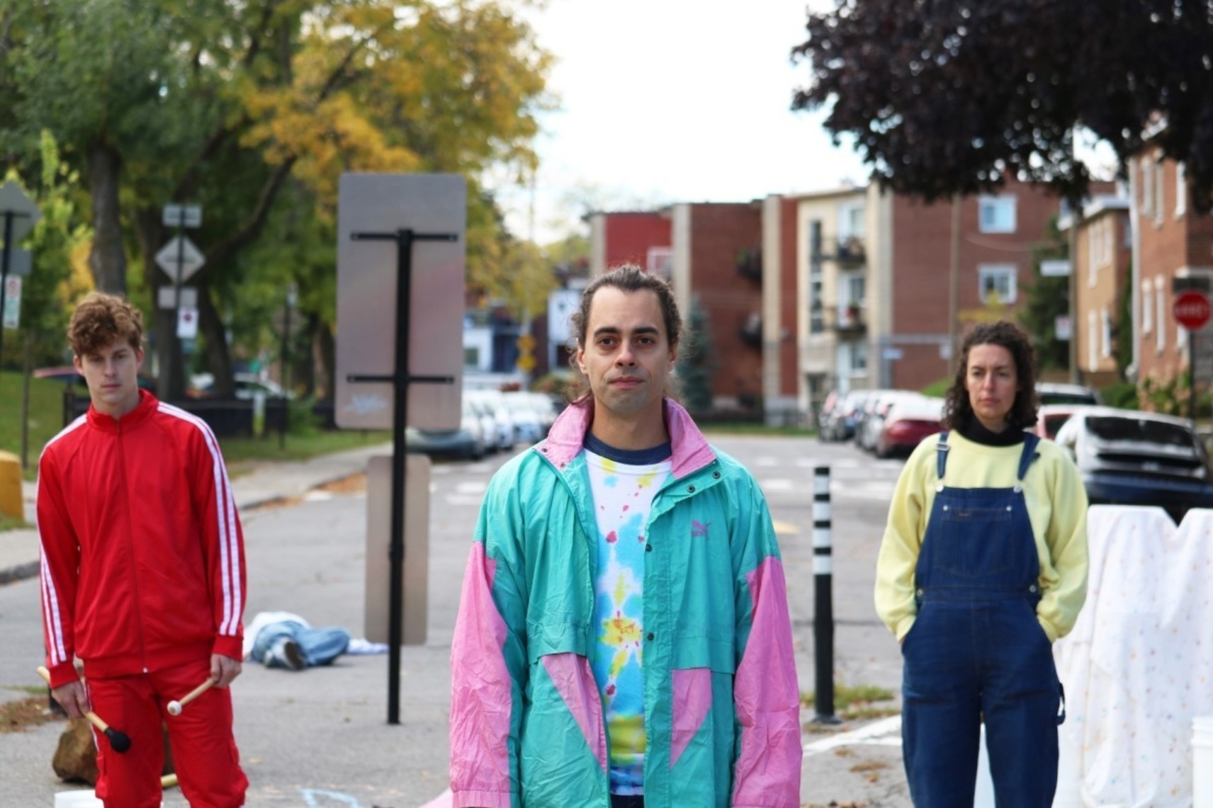 Photo en extérieur urbain de trois personnages. En avant-plan, un comédien portant un survêtement turquoise et rose. Derrière lui, un comédien en habit de sport rouge à gauche et une comédienne en salopette bleue et chandail jaune à droite.