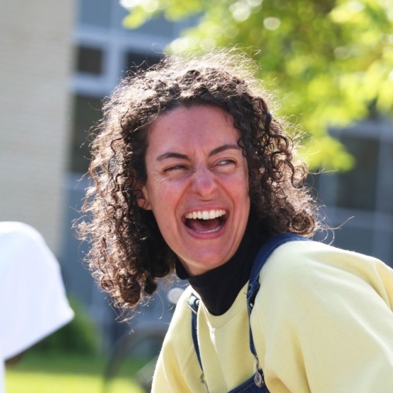 Photo en extérieur du personnage d'Amarelinha. Elle porte un haut jaune et regarde vers la droite, derrière elle, les yeux plissés en souriant.