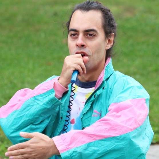 Photo en extérieur du personnage d'Ulysse, portant un survêtement turquoise et rose. Ses cheveux sont attachés et il parle dans un micro-jouet.