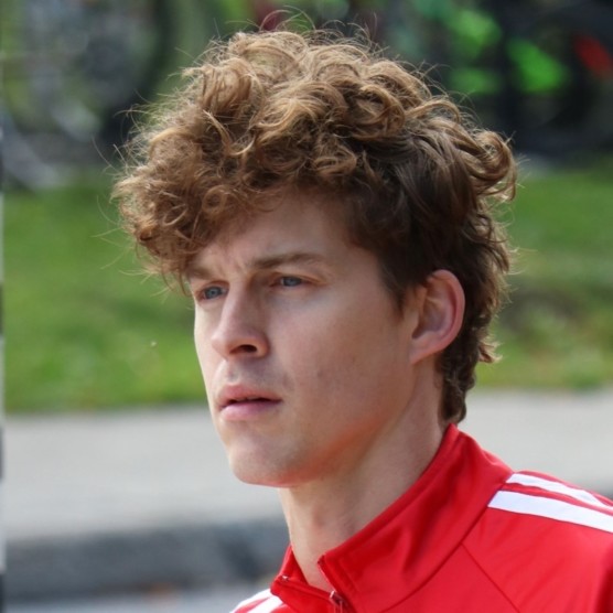 Photo en extérieur du personnage de Victor, portant un habit sportif rouge avec des lignes blanches. Ses yeux sont plissés dû à la lumière, il est légèrement de profil vers la gauche et il regarde devant lui.