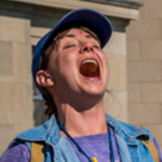 Photo en extérieur du personnage de Miranda, les yeux fermés et la bouche ouverte, la tête penchée vers l'arrière. Elle porte une casquette, un t-shirt coloré et une veste en jeans bleu.