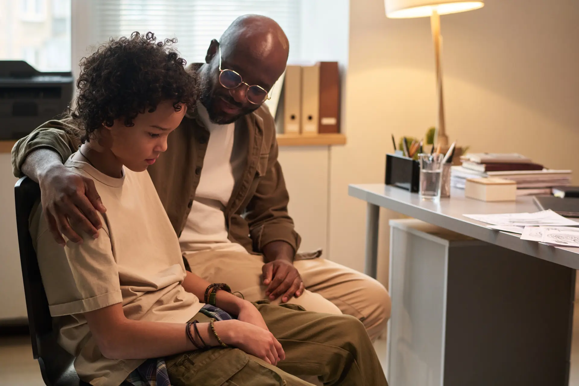 Young man embracing his upset son and looking at him while both sitting in school psychologist office at session