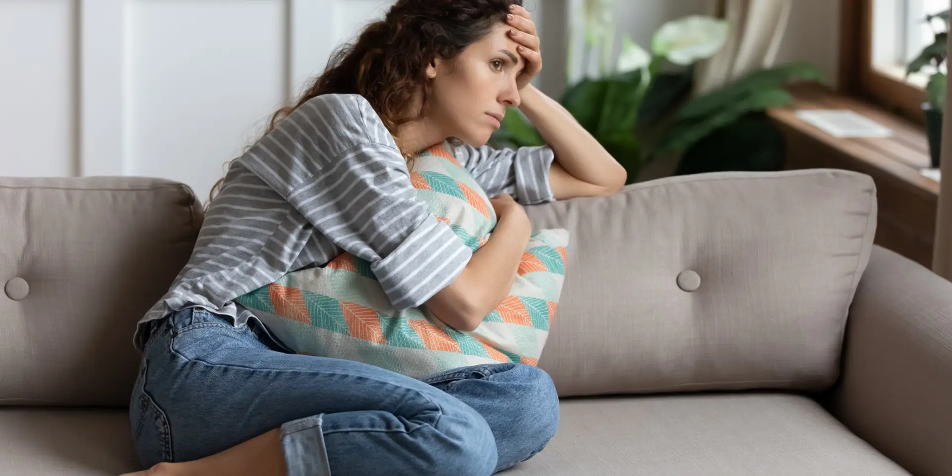 Frustrated young lady sitting on sofa, cuddling pillow, looking away at window. Lost in thoughts unhappy stressed millennial woman regretting of wrong decision, spending time alone in living room.